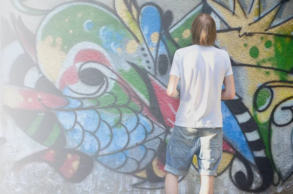 Photo of a young guy in denim shorts and a white shirt. The guy draws on the graffiti wall a drawing with aerosol paints of various colors. The concept of hooliganism and damage to property