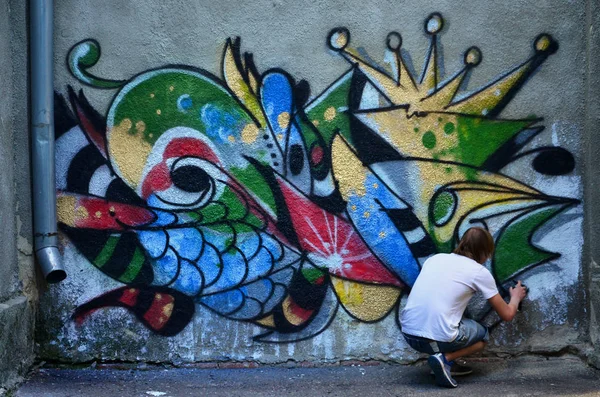 Photo in the process of drawing a graffiti pattern on an old concrete wall. Young long-haired blond guy draws an abstract drawing of different colors. Street art and vandalism concept