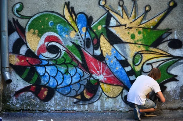 Photo of a young guy in denim shorts and a white shirt. The guy draws on the graffiti wall a drawing with aerosol paints of various colors. The concept of hooliganism and damage to property