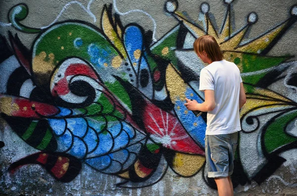 Photo of a young guy in denim shorts and a white shirt. The guy draws on the graffiti wall a drawing with aerosol paints of various colors. The concept of hooliganism and damage to property