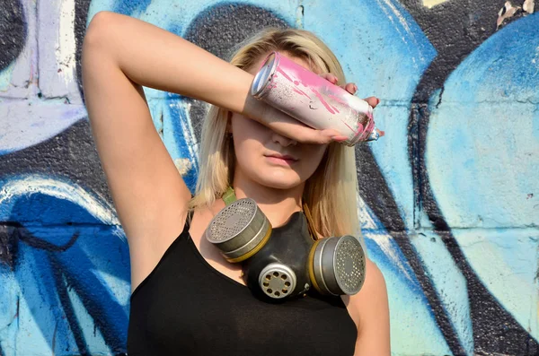 Young and beautiful sexy girl graffiti artist with gas mask on her neck hiding his eyes with a spray can standing on a wall background with a graffiti pattern in blue and purple tones