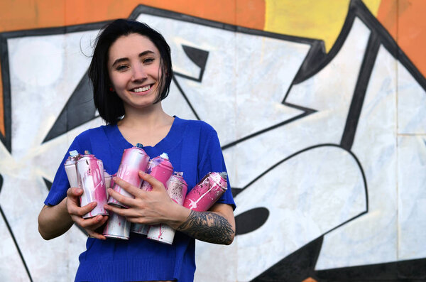 Портрет эмоциональной девушки с черными волосами и пирсингом. Фото девушки с канистрами аэрозольной краски в руках на фоне стены граффити. Концепция уличного искусства и использования аэрозольных красок
