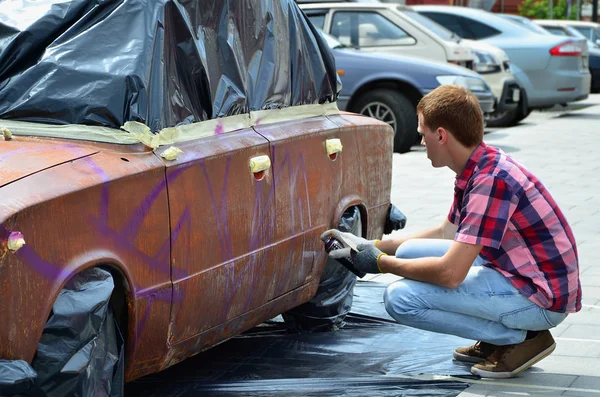 A young red-haired graffiti artist paints a new colorful graffiti on the car. Photo of the process of drawing a graffiti on a car close-up. The concept of street art and illegal vandalism