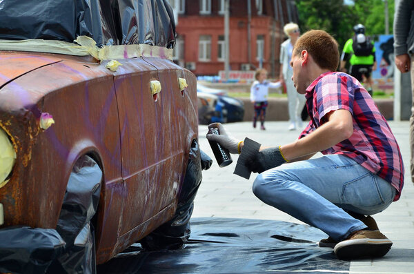 Молодой рыжеволосый граффити-художник рисует на автомобиле новые красочные граффити. Фотография процесса нанесения граффити на крупный план автомобиля. Концепция уличного искусства и незаконного вандализма
