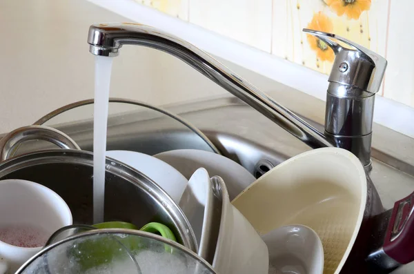 Dirty dishes and unwashed kitchen appliances lie in foam water under a tap from a kitchen faucet