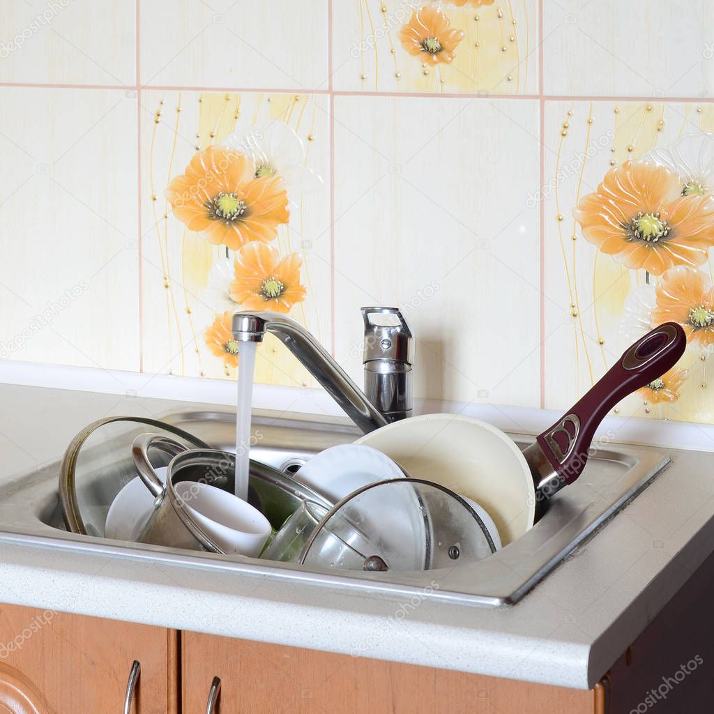 Dirty dishes and unwashed kitchen appliances lie in foam water under a tap from a kitchen faucet