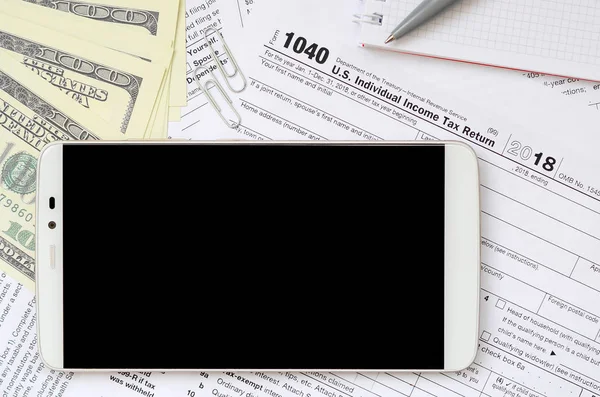 Composition of items lying on the 1040 tax form. Dollar bills, pen, smartphone, paper clip and notepad. Clean black screen smartphone for text placement