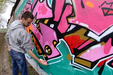 Gri kapüşonlu genç bir adam, yağmurlu havalarda grafiti bir duvar pembe ve yeşil renklerde boyar. Balıkgözü atış