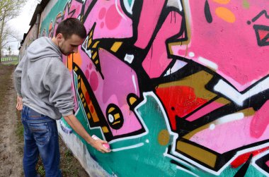 Gri kapüşonlu genç bir adam, yağmurlu havalarda grafiti bir duvar pembe ve yeşil renklerde boyar. Balıkgözü atış