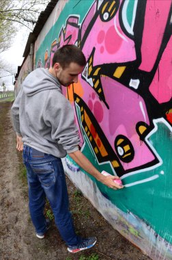 Gri kapüşonlu genç bir adam, yağmurlu havalarda grafiti bir duvar pembe ve yeşil renklerde boyar. Balıkgözü atış