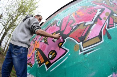 Gri kapüşonlu genç bir adam, yağmurlu havalarda grafiti bir duvar pembe ve yeşil renklerde boyar. Balıkgözü atış