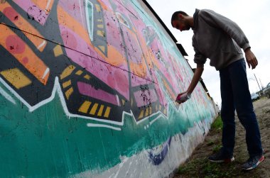 Gri kapüşonlu genç bir adam, yağmurlu havalarda grafiti bir duvar pembe ve yeşil renklerde boyar. Balıkgözü atış