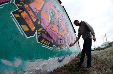 Gri kapüşonlu genç bir adam, yağmurlu havalarda grafiti bir duvar pembe ve yeşil renklerde boyar. Balıkgözü atış