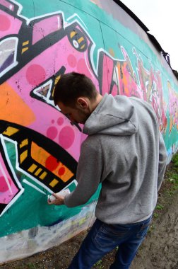 Gri kapüşonlu genç bir adam, yağmurlu havalarda grafiti bir duvar pembe ve yeşil renklerde boyar. Balıkgözü atış
