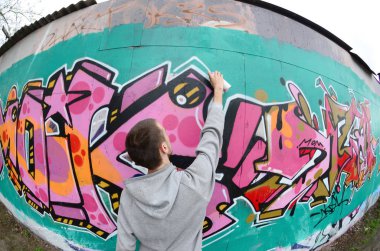 Gri kapüşonlu genç bir adam, yağmurlu havalarda grafiti bir duvar pembe ve yeşil renklerde boyar. Balıkgözü atış