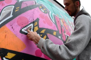 Gri kapüşonlu genç bir adam, yağmurlu havalarda grafiti bir duvar pembe ve yeşil renklerde boyar. Balıkgözü atış