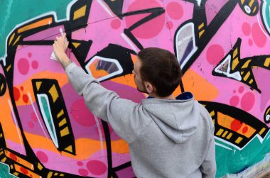 Gri kapüşonlu genç bir adam, yağmurlu havalarda grafiti bir duvar pembe ve yeşil renklerde boyar. Balıkgözü atış