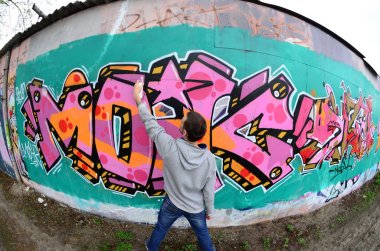 Gri kapüşonlu genç bir adam, yağmurlu havalarda grafiti bir duvar pembe ve yeşil renklerde boyar. Balıkgözü atış