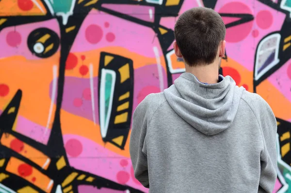 A young graffiti artist in a gray hoodie looks at the wall with his graffiti in pink and green colors on a wall in rainy weather. Street art concept