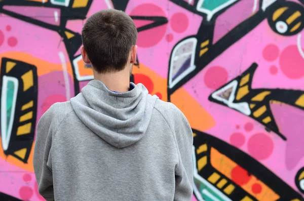 A young graffiti artist in a gray hoodie looks at the wall with his graffiti in pink and green colors on a wall in rainy weather. Street art concept