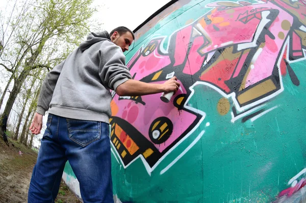 A young guy in a gray hoodie paints graffiti in pink and green colors on a wall in rainy weather. Fisheye shot