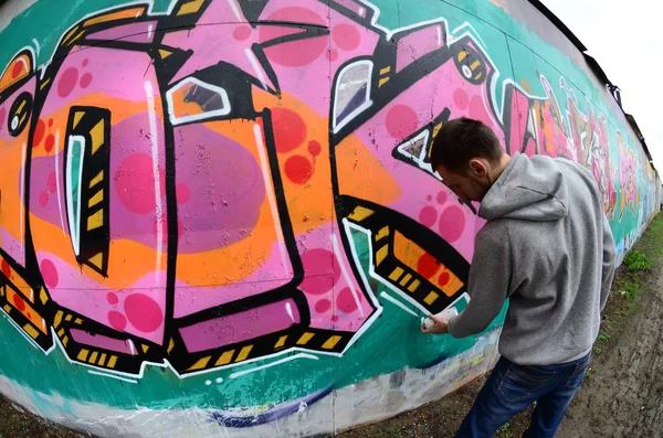 一个穿着灰色帽衫的年轻人在雨天的墙上画着粉红色和绿色的涂鸦 鱼眼射击 — 图库照片