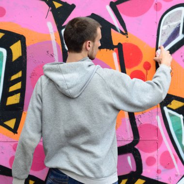 Gri kapüşonlu genç bir adam grafiti bir duvar pembe ve yeşil renklerde yağmurlu havalarda boyar.