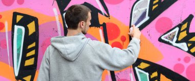 Gri kapüşonlu genç bir adam grafiti bir duvar pembe ve yeşil renklerde yağmurlu havalarda boyar.