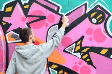 Gri kapüşonlu genç bir adam grafiti bir duvar pembe ve yeşil renklerde yağmurlu havalarda boyar.