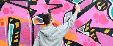 Gri kapüşonlu genç bir adam grafiti bir duvar pembe ve yeşil renklerde yağmurlu havalarda boyar.