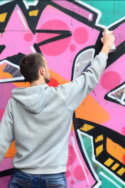 Gri kapüşonlu genç bir adam grafiti bir duvar pembe ve yeşil renklerde yağmurlu havalarda boyar.