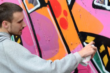 Gri kapüşonlu genç bir adam grafiti bir duvar pembe ve yeşil renklerde yağmurlu havalarda boyar.