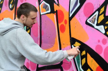 Gri kapüşonlu genç bir adam grafiti bir duvar pembe ve yeşil renklerde yağmurlu havalarda boyar.
