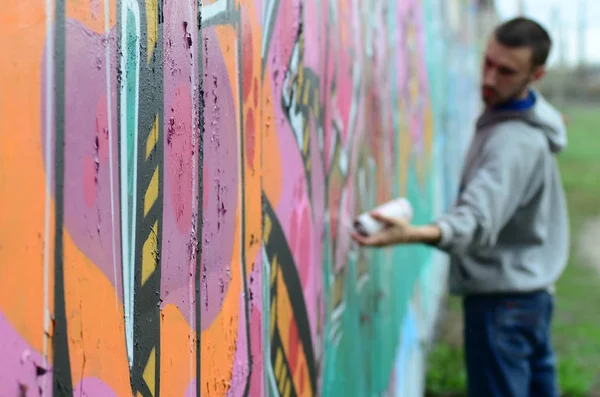 Young guy in a gray hoodie paints graffiti in pink and green colors on a wall in rainy weather Focus on the fragment of wall and blurred artist