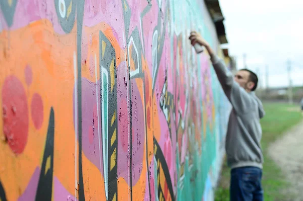 Young guy in a gray hoodie paints graffiti in pink and green colors on a wall in rainy weather Focus on the fragment of wall and blurred artist