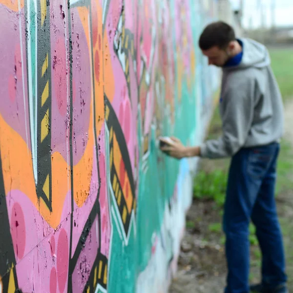 Young guy in a gray hoodie paints graffiti in pink and green colors on a wall in rainy weather Focus on the fragment of wall and blurred artist