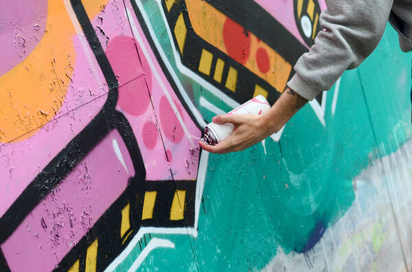 Hand of a young guy in a gray hoodie paints graffiti in pink and green colors on a wall in rainy weather