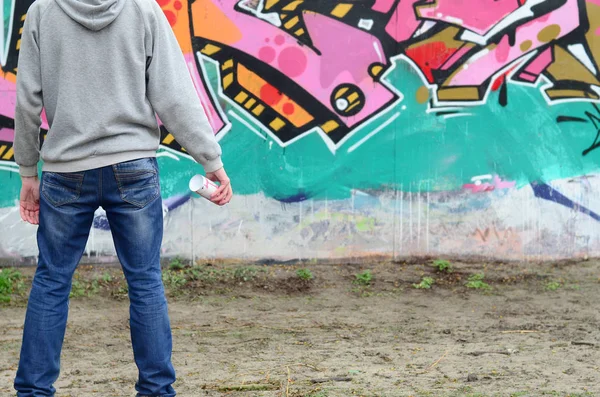 A young graffiti artist in a gray hoodie looks at the wall with his graffiti in pink and green colors on a wall in rainy weather. Street art concept