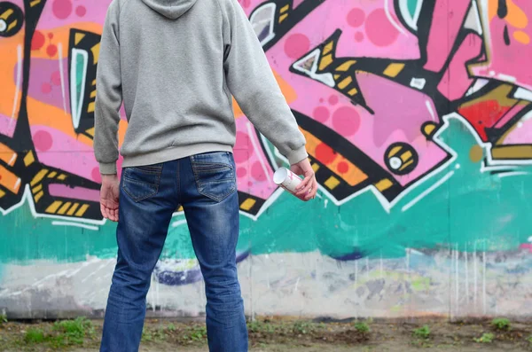 A young graffiti artist in a gray hoodie looks at the wall with his graffiti in pink and green colors on a wall in rainy weather. Street art concept