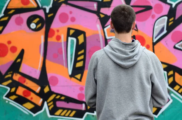 A young graffiti artist in a gray hoodie looks at the wall with his graffiti in pink and green colors on a wall in rainy weather. Street art concept