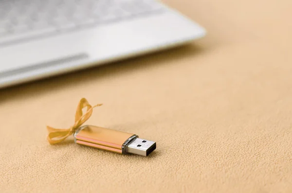 Orange usb flash memory card with a bow lies on a blanket of soft and furry light orange fleece fabric beside to a white laptop. Classic female gift design for a memory card