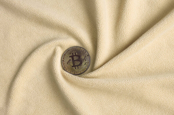 The golden bitcoin lies on a blanket made of soft and fluffy light orange fleece fabric with a large number of relief folds. The shape of the folds resembles a fan from a video card cooler