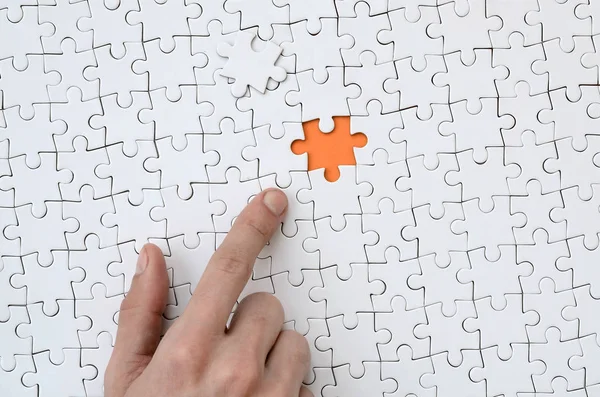 The texture of a white jigsaw puzzle in the assembled state with one missing element, forming an orange space, pointed to by the finger of the male hand