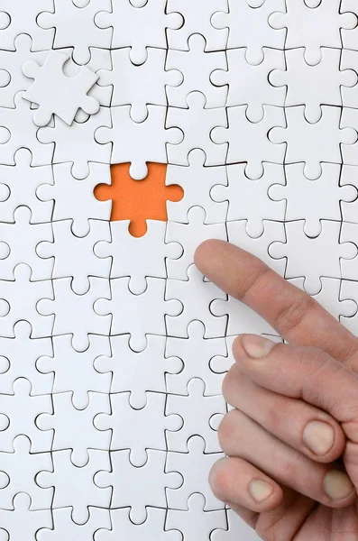 La trama di un puzzle bianco nello stato assemblato con un elemento mancante, formando uno spazio arancione, indicato dal dito della mano maschile — Foto Stock
