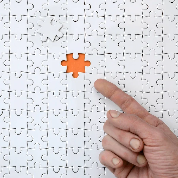 The texture of a white jigsaw puzzle in the assembled state with one missing element, forming an orange space, pointed to by the finger of the male hand
