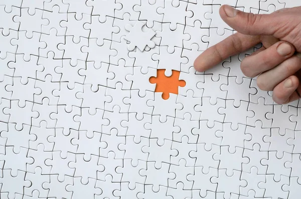 The texture of a white jigsaw puzzle in the assembled state with one missing element, forming an orange space, pointed to by the finger of the male hand