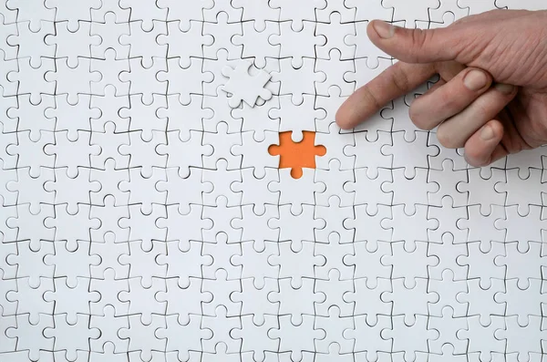 The texture of a white jigsaw puzzle in the assembled state with one missing element, forming an orange space, pointed to by the finger of the male hand