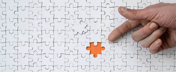 The texture of a white jigsaw puzzle in the assembled state with one missing element, forming an orange space, pointed to by the finger of the male hand
