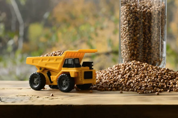 A small yellow toy truck is loaded with brown grains of buckwheat around the buckwheat pile and a glass of croup. A car on a wooden surface against a background of autumn forest. Buckwheat delivery