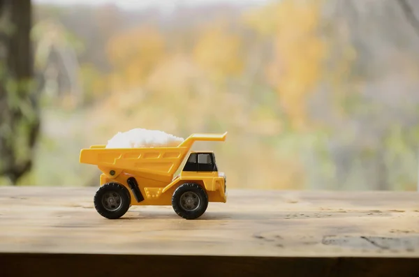 A small yellow toy truck is loaded with a stone of white salt. A car on a wooden surface against a background of autumn forest. Extraction and transportation of salt